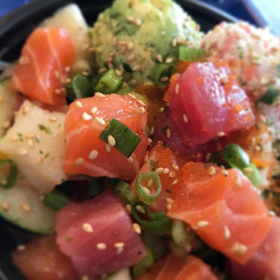 Close-up of a Poki Bowl with salmon, tuna, avocado, and green onions, sprinkled with sesame seeds.